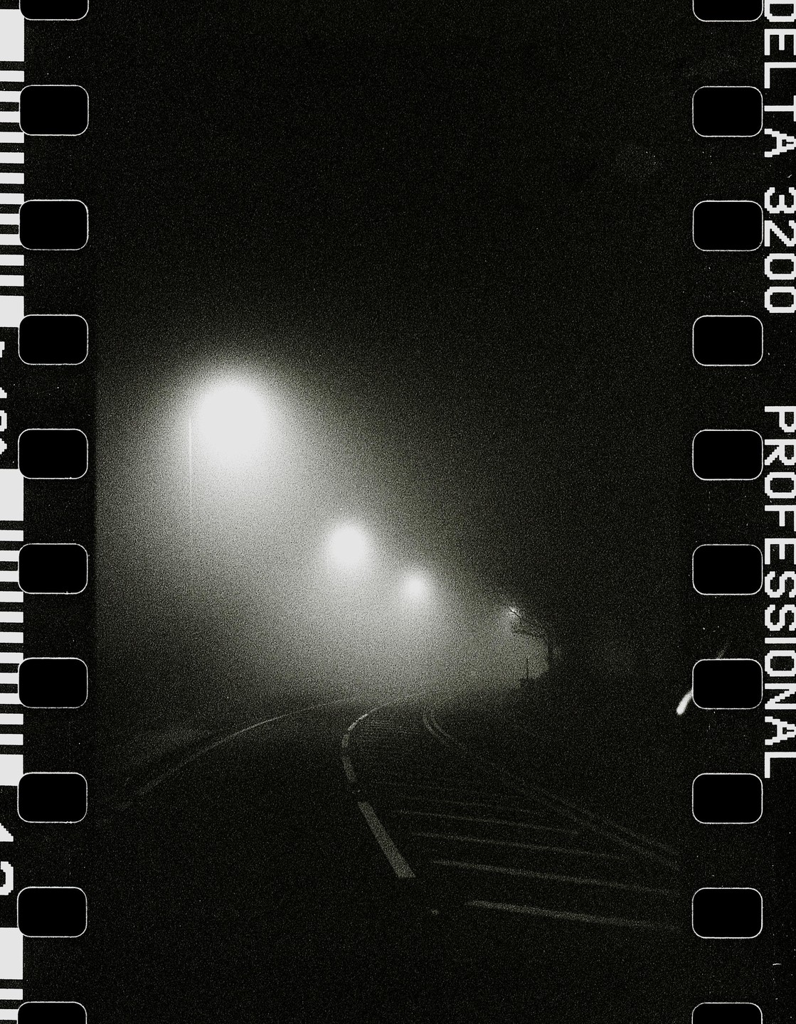 A winding road is illuminated by two streetlights in the fog at night.