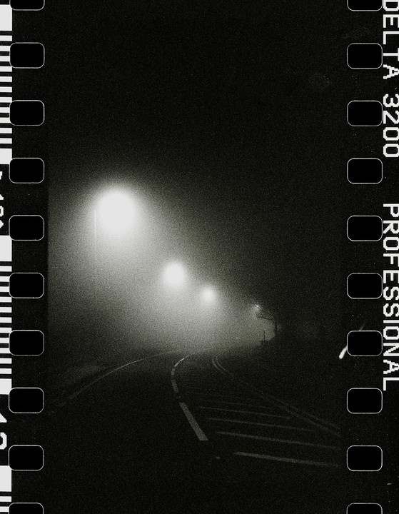 A winding road is illuminated by two streetlights in the fog at night.