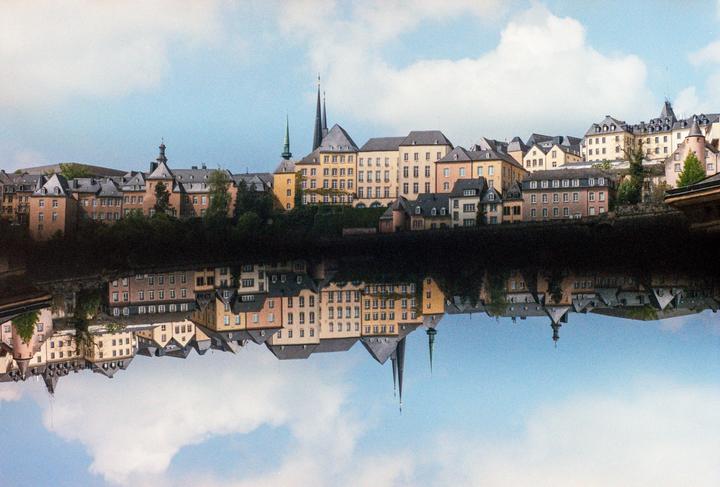 A city with many buildings and spires is reflected in the water under a cloudy sky.