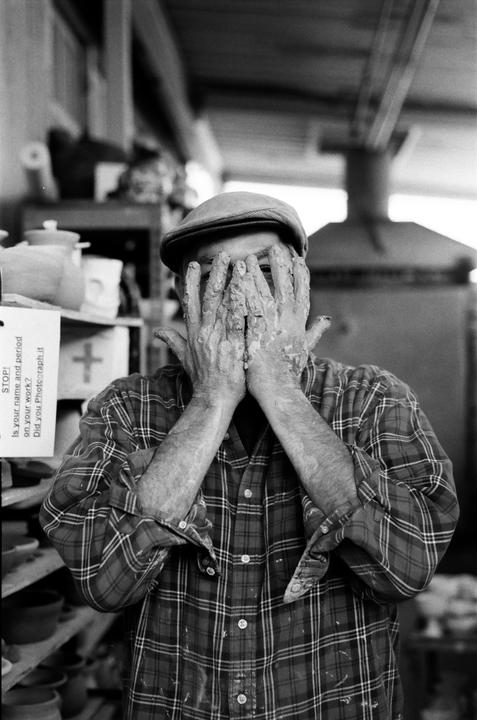 A man wearing a flat cap and plaid shirt covers his face with his clay-covered hands.