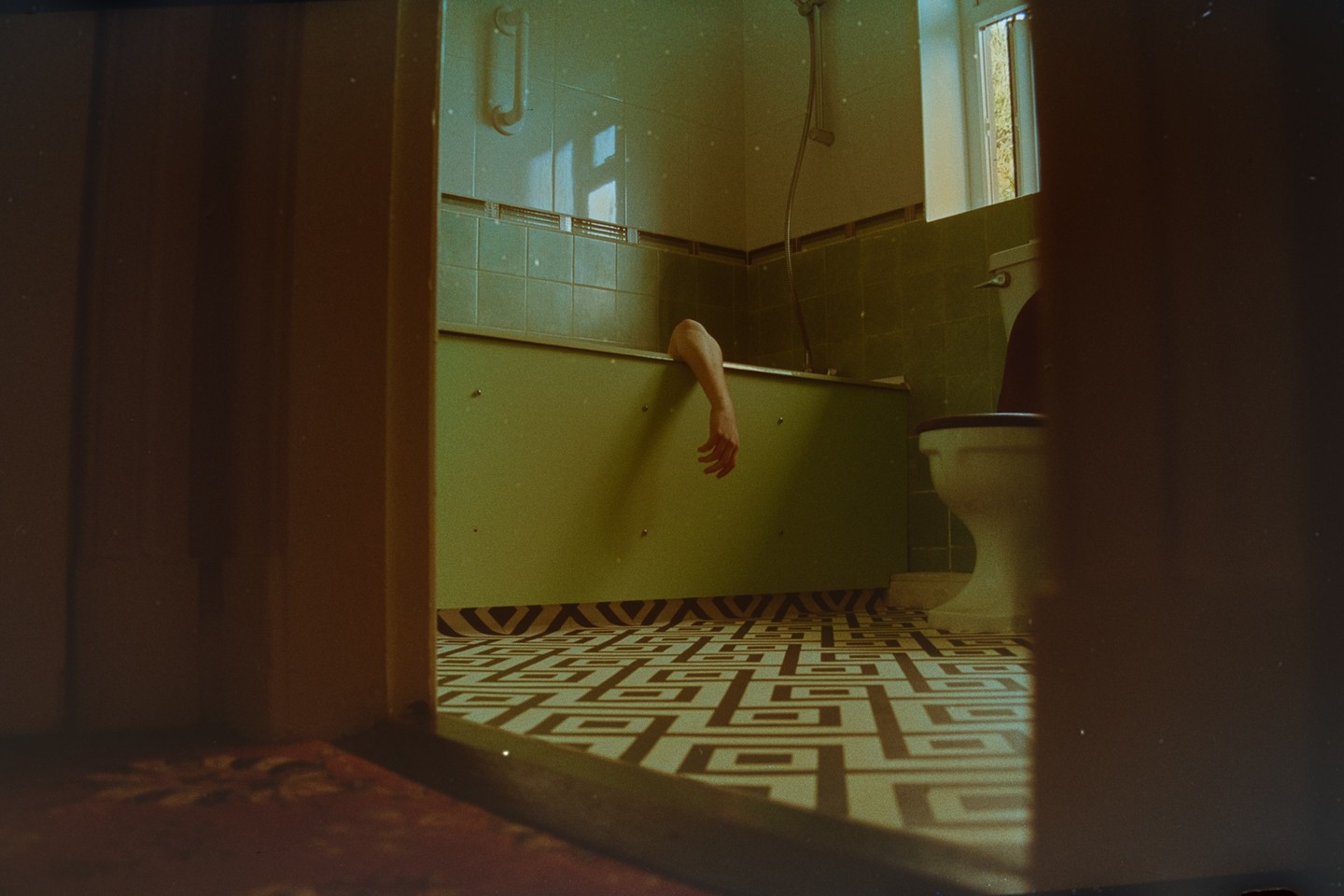 An arm hangs out of a bathtub in a green-tiled bathroom with patterned floor tiles, viewed through a doorway.