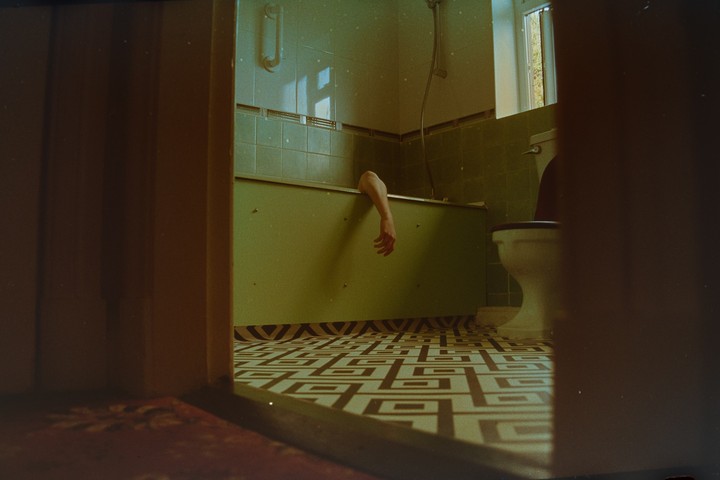 An arm hangs out of a bathtub in a green-tiled bathroom with patterned floor tiles, viewed through a doorway.