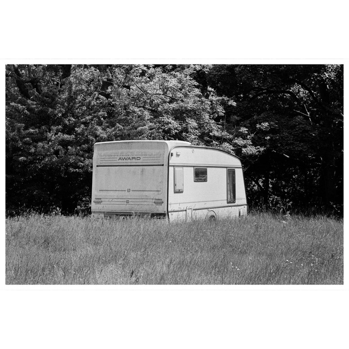 An abandoned caravan sits in a field of tall grass with trees in the background.