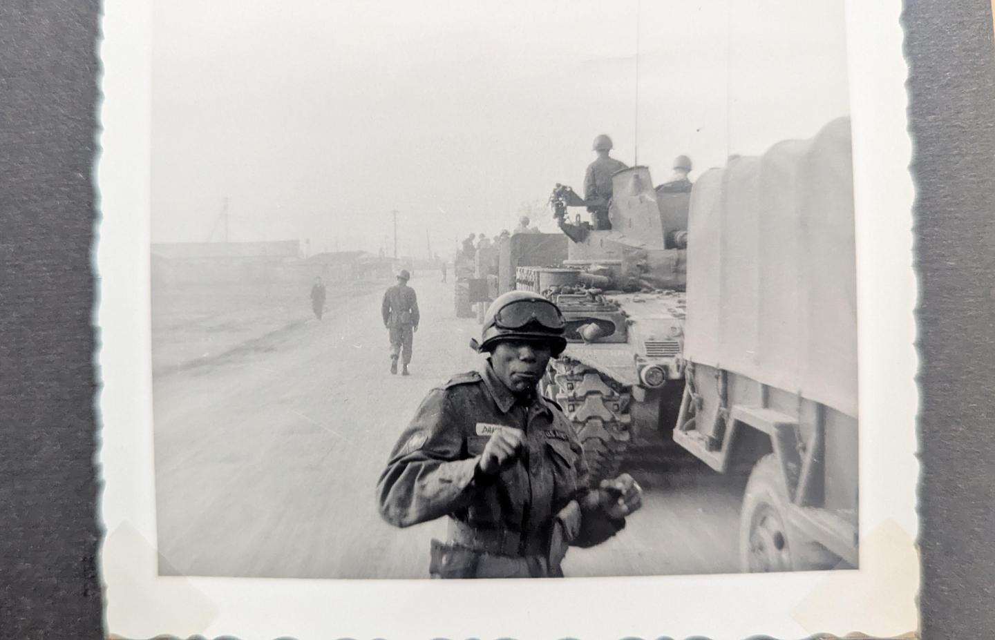 A man in military uniform and goggles stands in the foreground with military vehicles and other soldiers behind him on a hazy day.