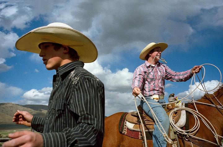 Two cowboys wear hats, one on horseback holding a rope and the other standing with a mountain in the background.