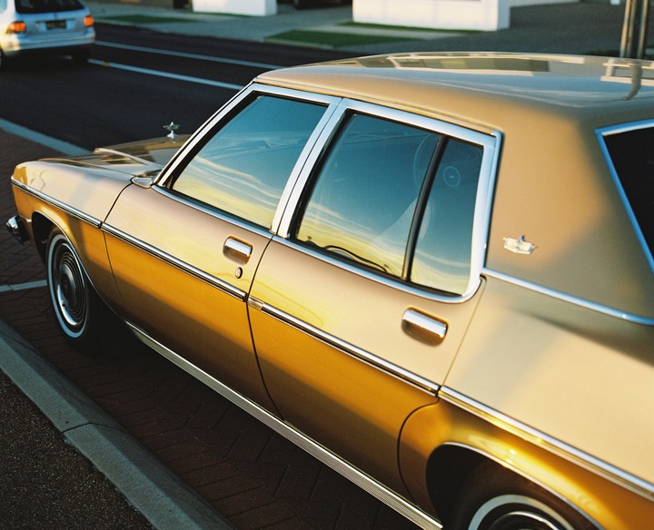 A gold vintage car is parked on a brick curb with reflections of the sunset in its windows.