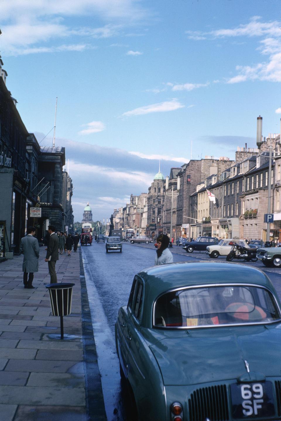 A street scene shows a woman standing next to a parked car, with other cars and people on the street and buildings lining both sides.
