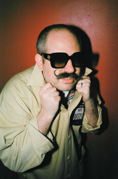 A man with a handlebar mustache and sunglasses holds his collar up against a red wall.