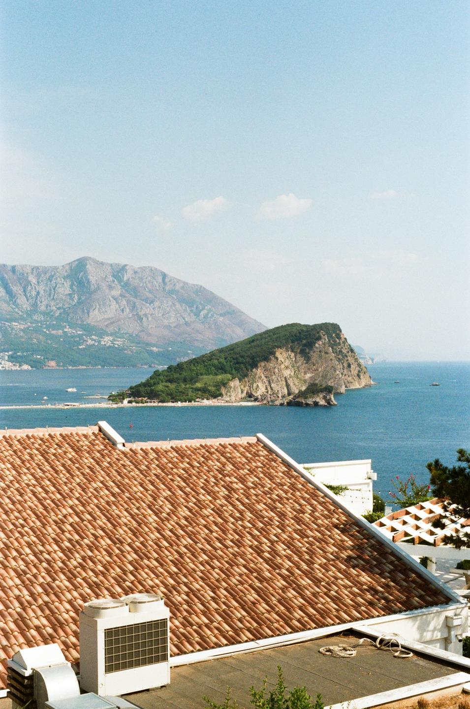 A tiled rooftop overlooks a bay with a rocky island and mountains in the distance.