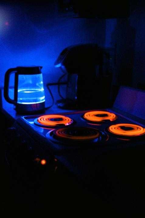 A blue illuminated kettle and a coffee maker sit on a counter next to a stove with four glowing orange burners.