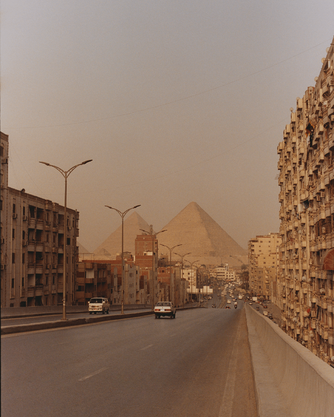A wide road with cars and buildings on either side leads towards the pyramids in the distance.