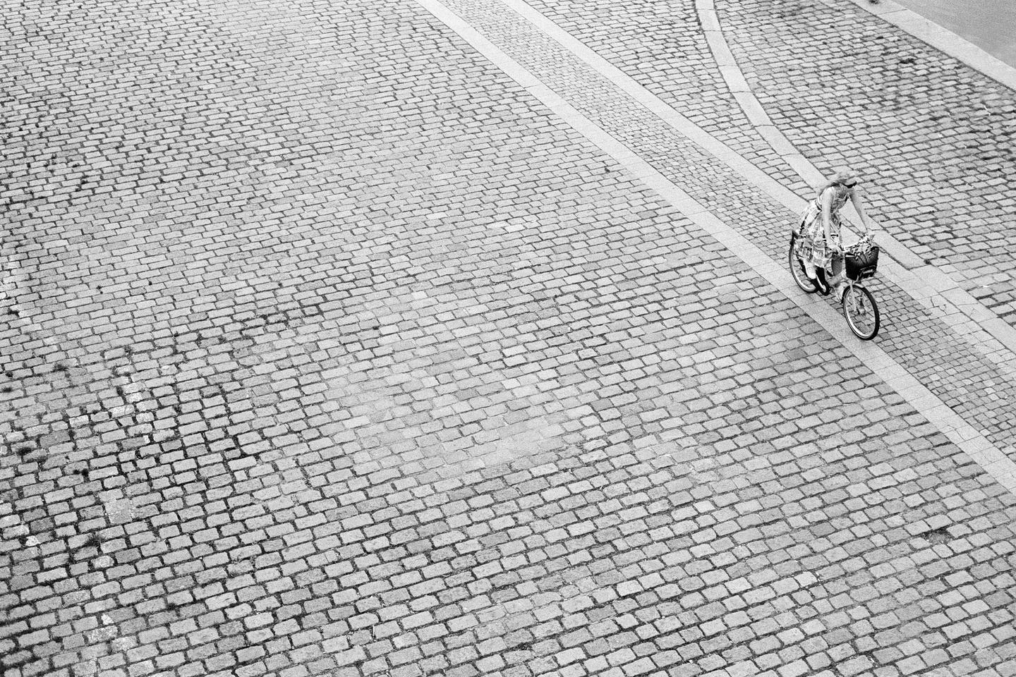 A woman rides a bicycle with a basket on a cobblestone street.