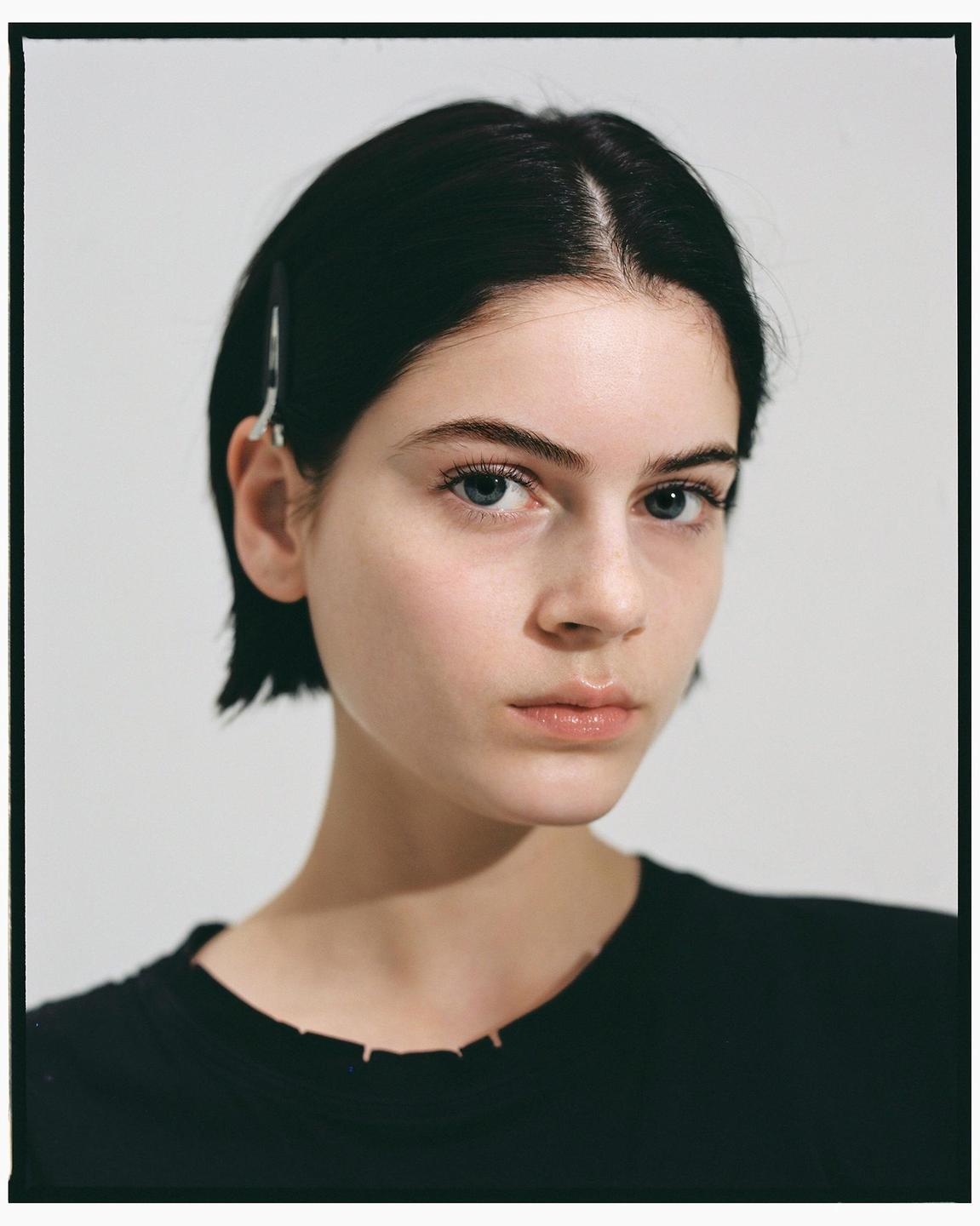 A woman with short dark hair and a black shirt looks directly at the viewer in a studio portrait.