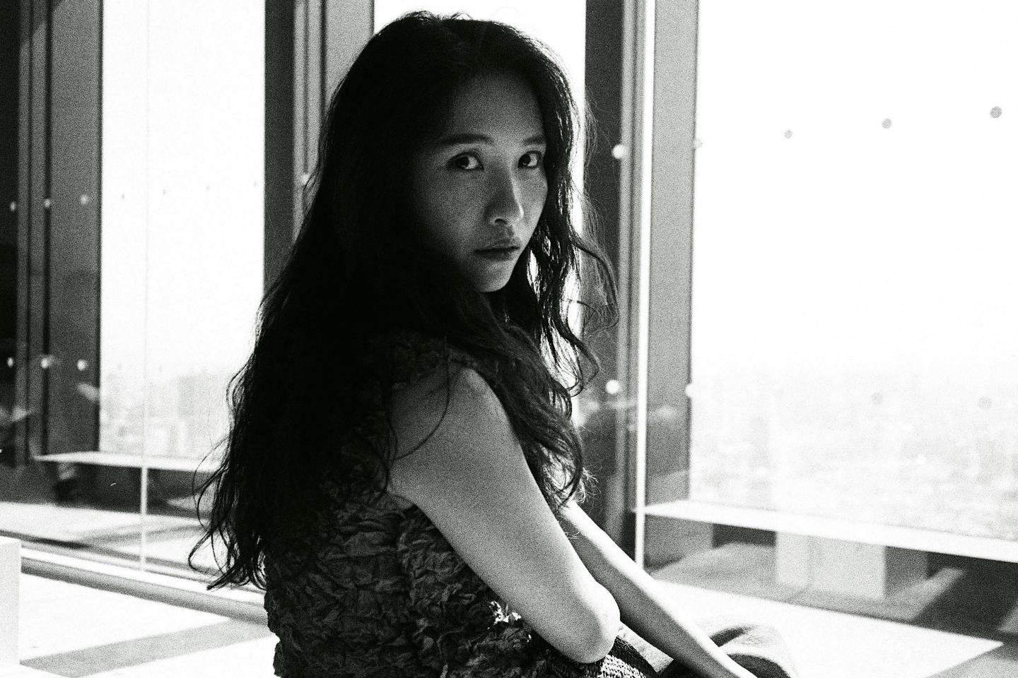 A woman with long dark hair looks over her shoulder at the viewer, sitting in front of a large window.