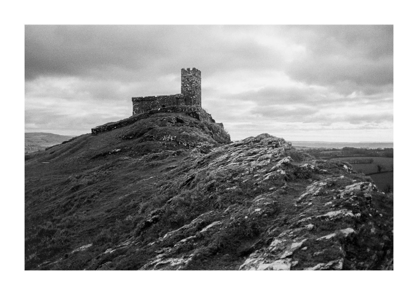 A stone church with a tall tower stands atop a grassy hill under a cloudy sky.