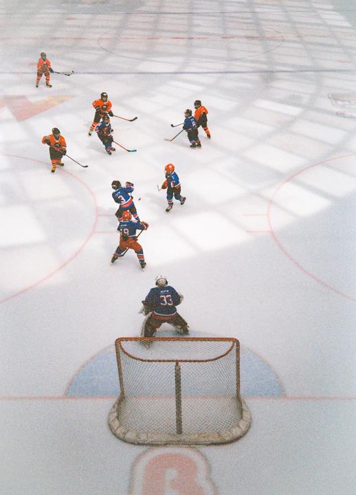 A high-angle view shows a hockey game in progress with players in orange and blue jerseys on an ice rink.