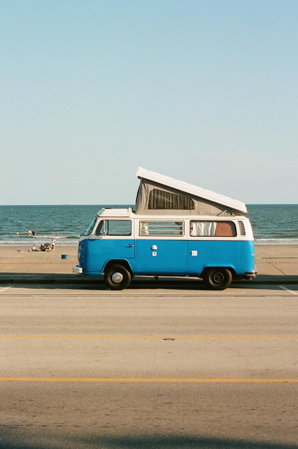 A blue and white camper van is parked on the side of a road next to a beach with people in the water.