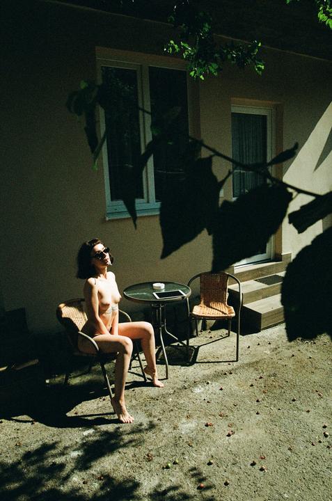 A woman sits topless in a chair on a patio next to a small table with a tablet and ashtray.