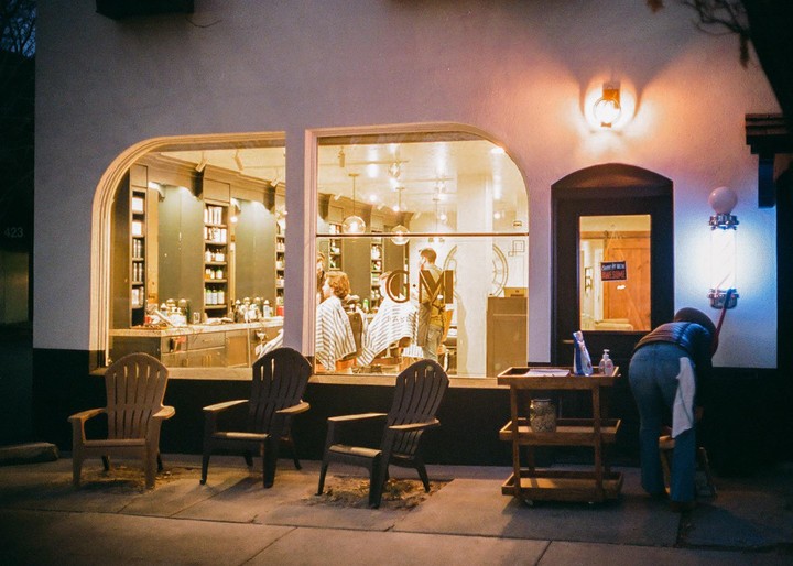 A person sweeps the sidewalk outside a barbershop at night, with customers visible through the window.