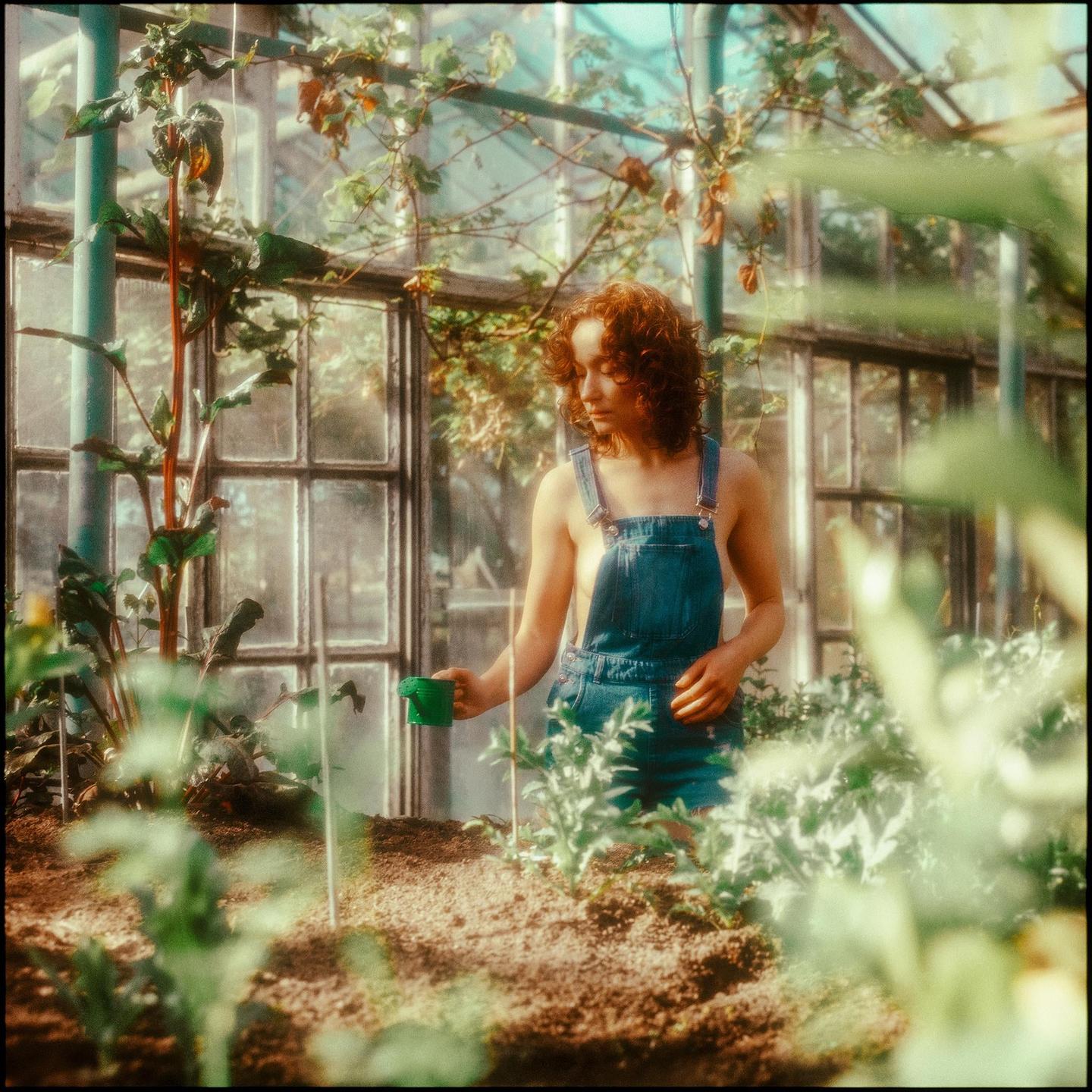 A woman in denim overalls holds a green watering can in a greenhouse.