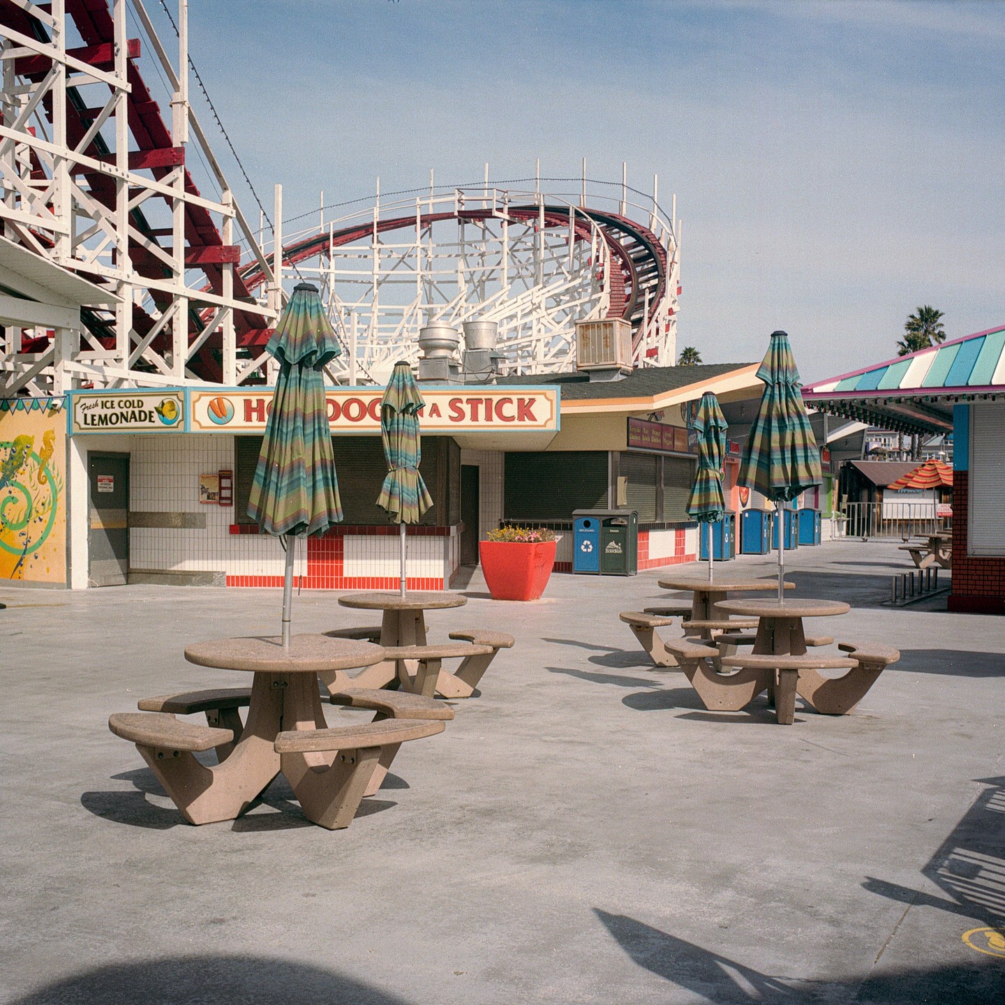 A boardwalk food stand with picnic tables and umbrellas sits in front of a roller coaster.