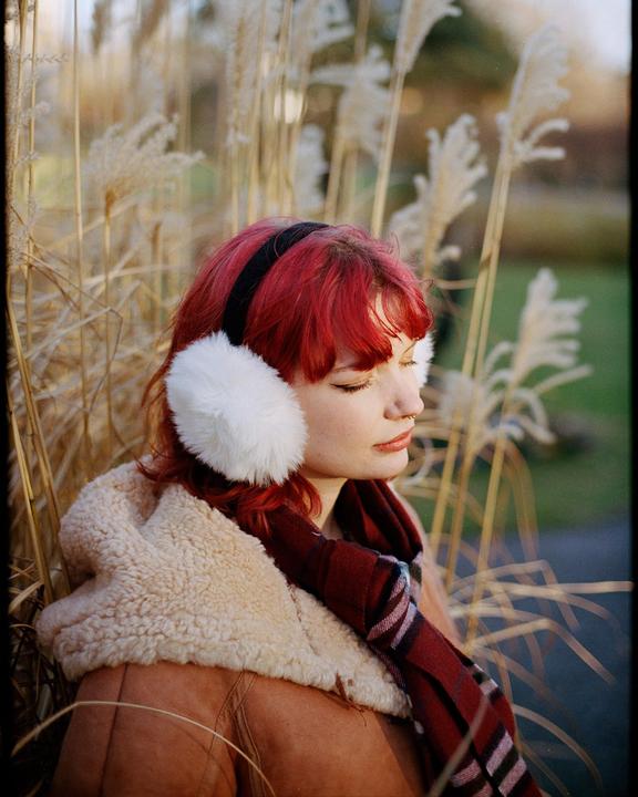 A woman with red hair and white earmuffs is sitting among tall grasses.