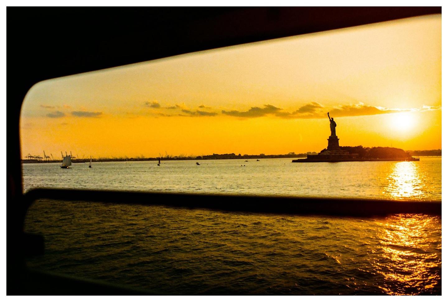 The Statue of Liberty is silhouetted against a bright orange sunset over the water, as seen through a window.