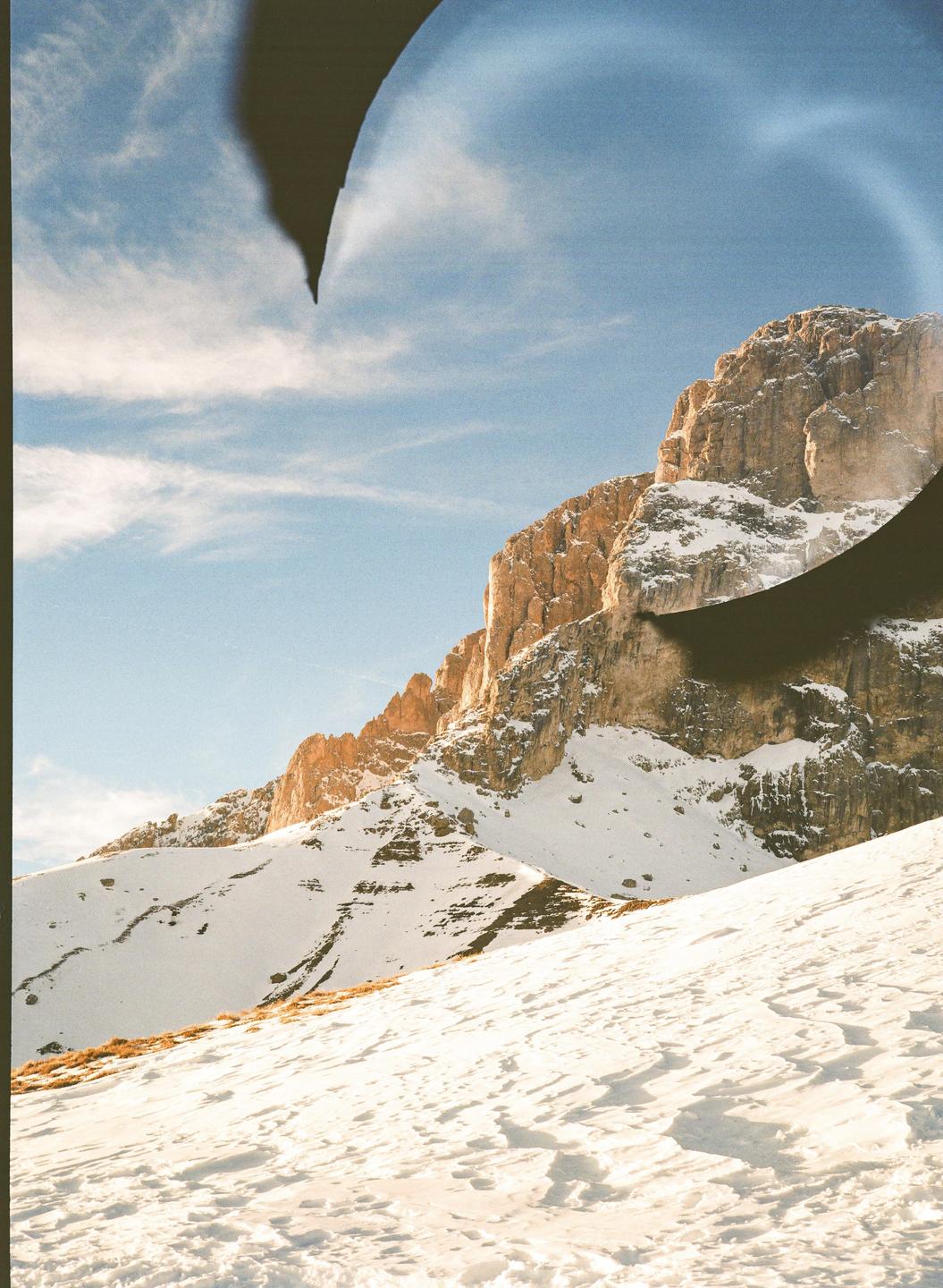 A snow-covered mountain slope leads up to a rocky mountain under a blue sky with some clouds.