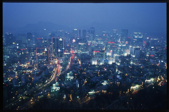 A city at night is illuminated by the lights of buildings and cars, with mountains visible in the distance.