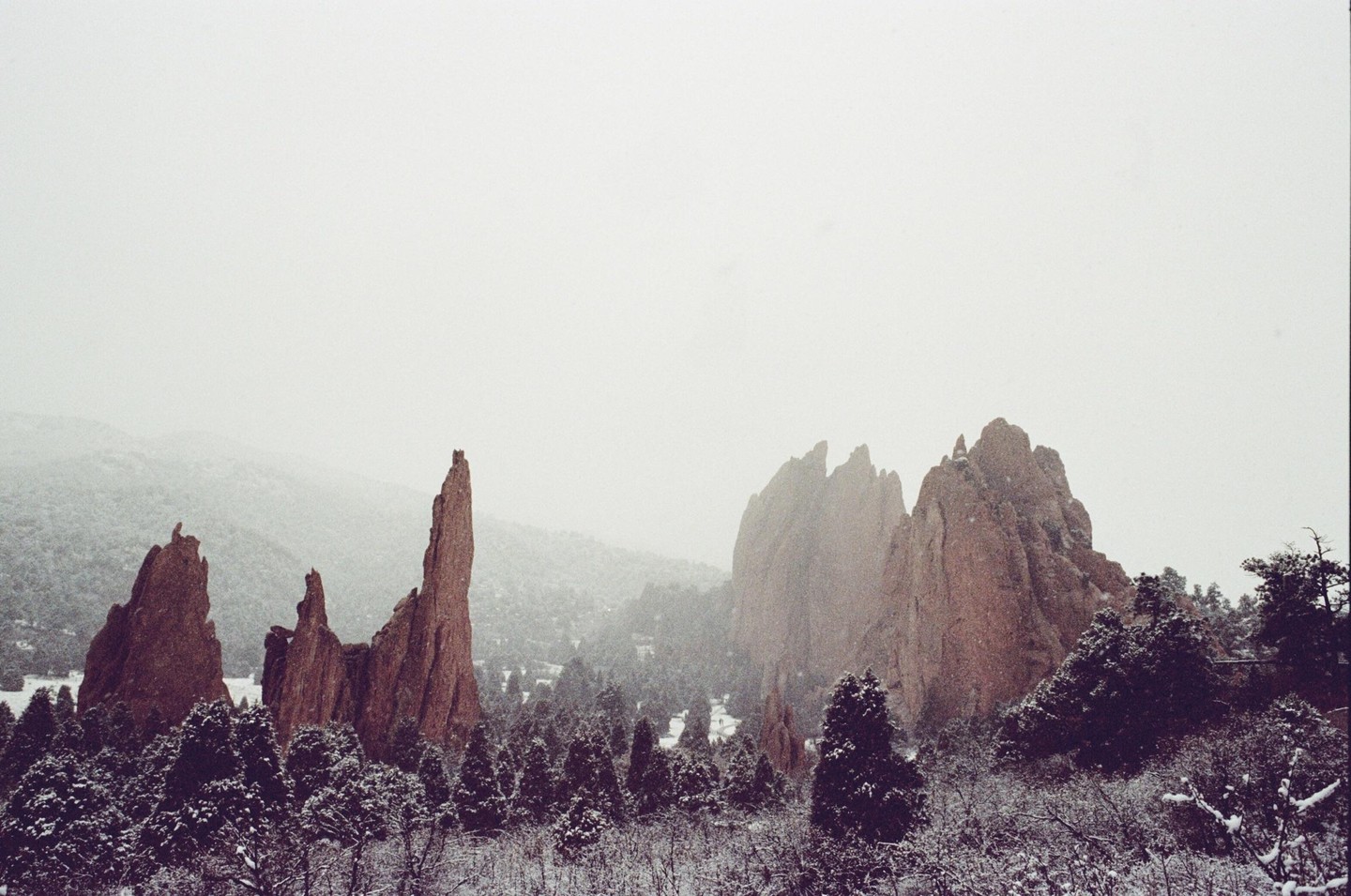 Red rock formations rise above a snow-covered forest under a hazy sky.