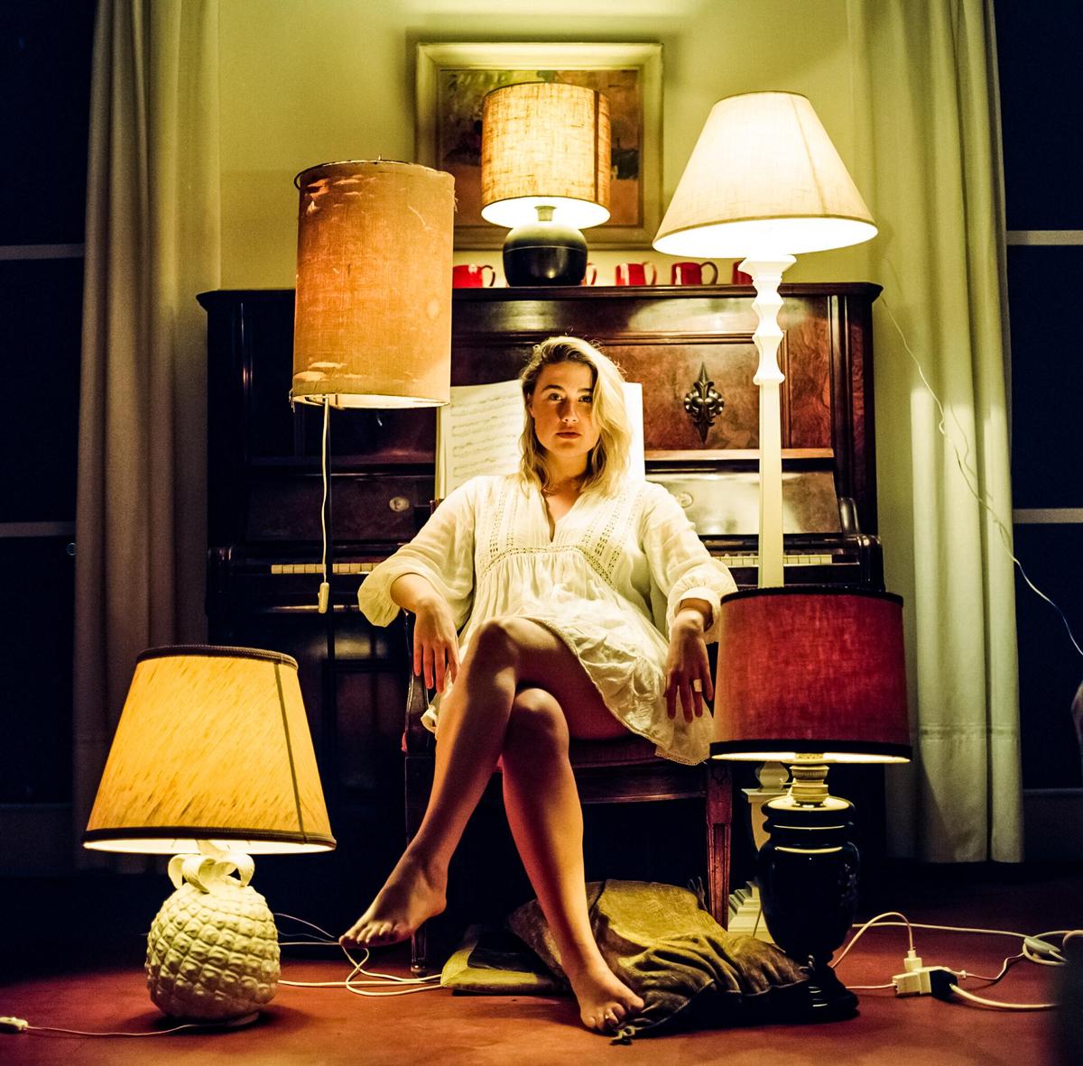 A woman sits in a chair in front of a piano, surrounded by lamps.