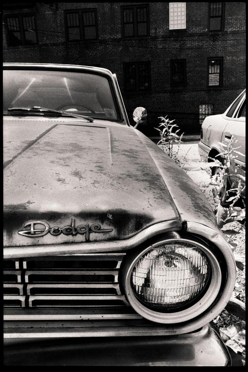 A close-up view of the front of an old Dodge car with a brick building in the background.
