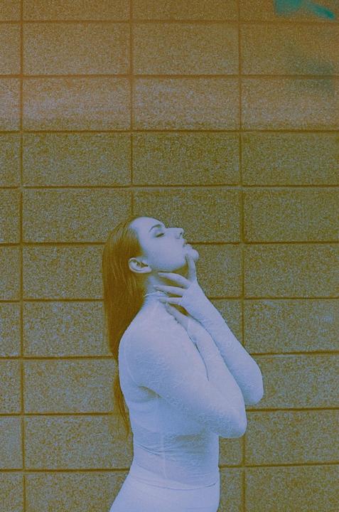 A woman in a white lace garment poses with her head tilted back and her hands on her neck against a textured wall.