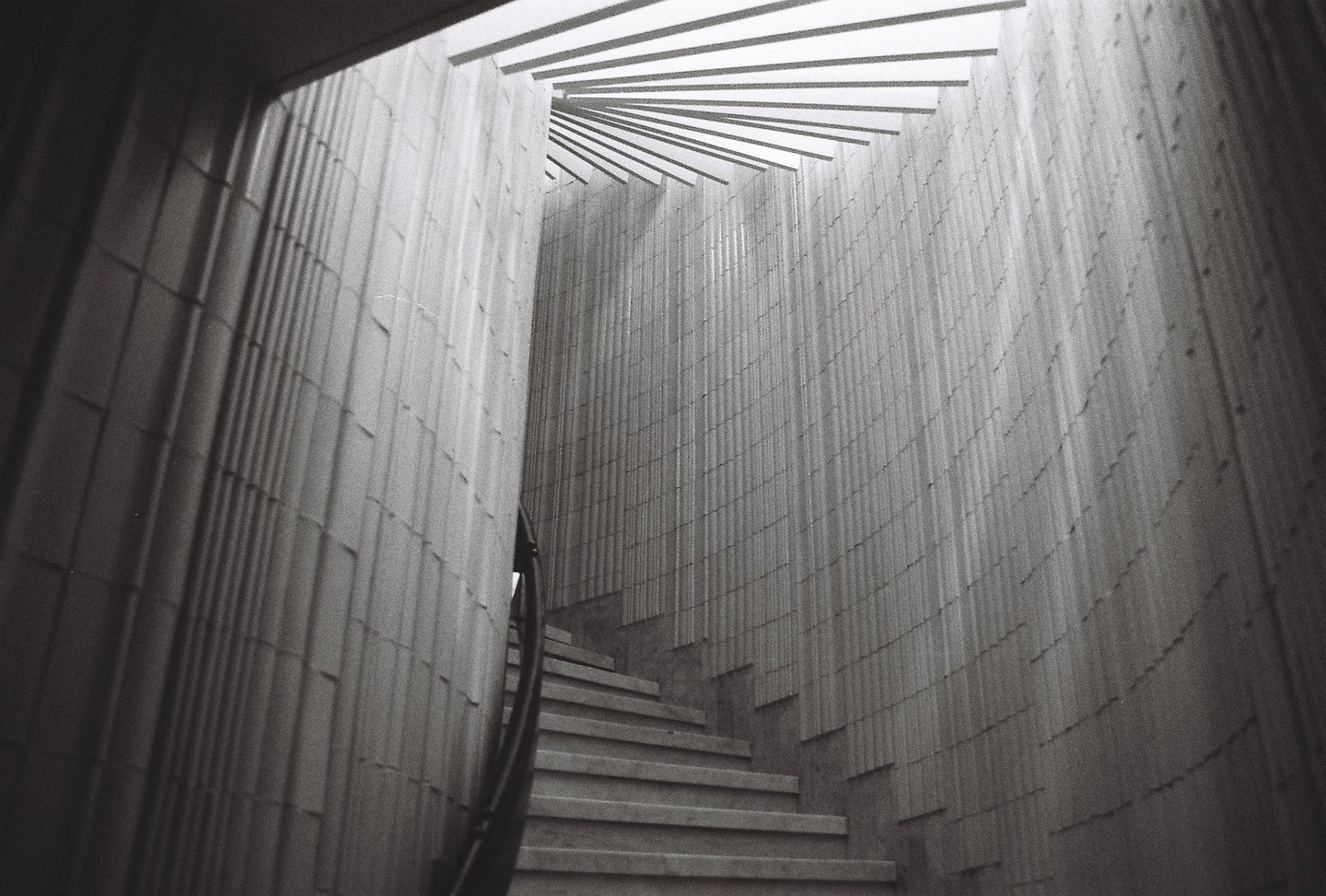 A narrow, winding staircase with a handrail ascends between two textured walls, leading towards a slatted ceiling.
