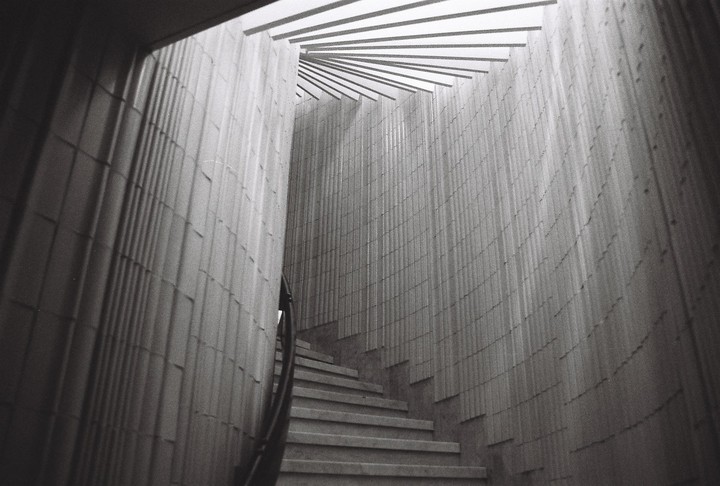 A narrow, winding staircase with a handrail ascends between two textured walls, leading towards a slatted ceiling.