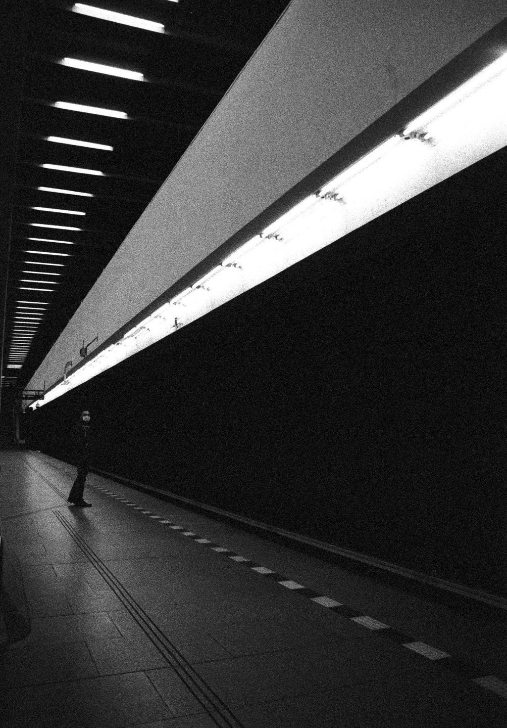 A person stands on a subway platform with bright fluorescent lights illuminating the ceiling and wall.