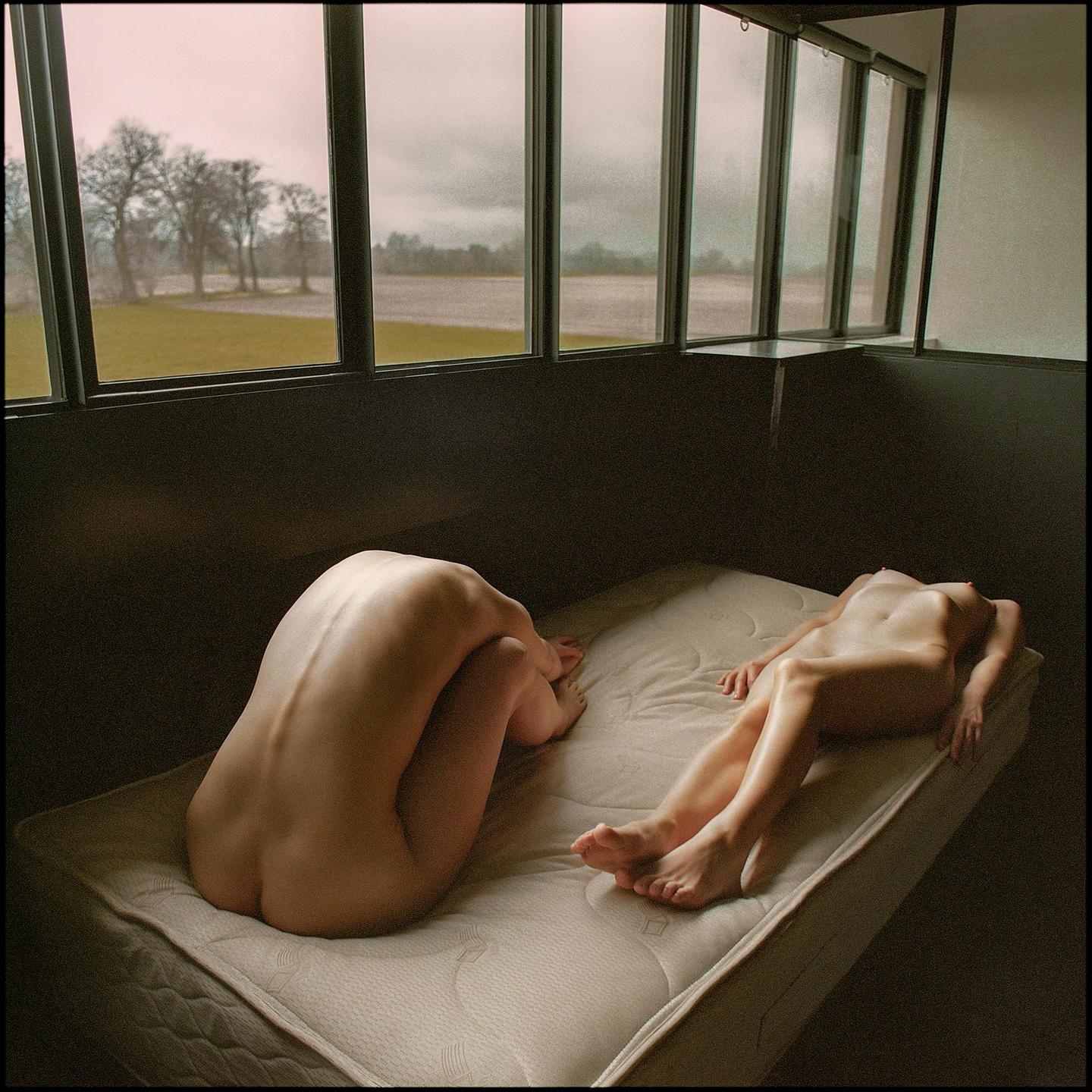 Two nude women are on a mattress in a room with large windows overlooking a field with trees.