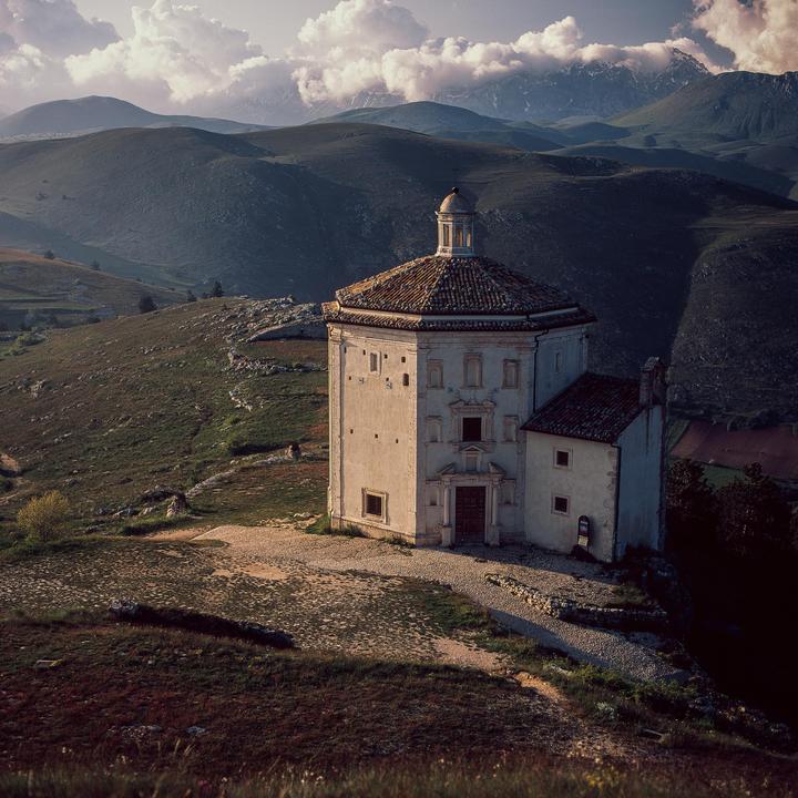 A small, octagonal church with a tiled roof stands on a grassy hill overlooking a valley with mountains in the distance.