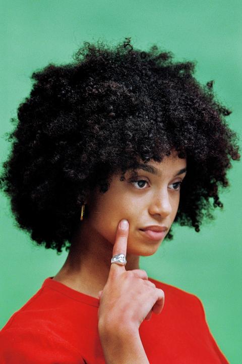 A woman with curly hair wears a red shirt and a ring while touching her cheek against a green background.