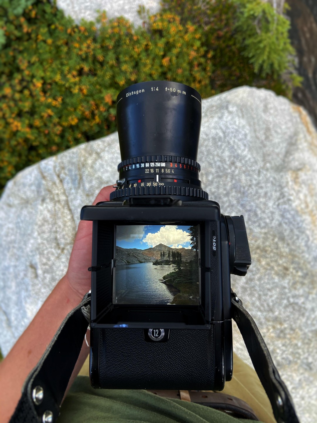 A person holds a camera with a lake and mountains visible in its viewfinder.