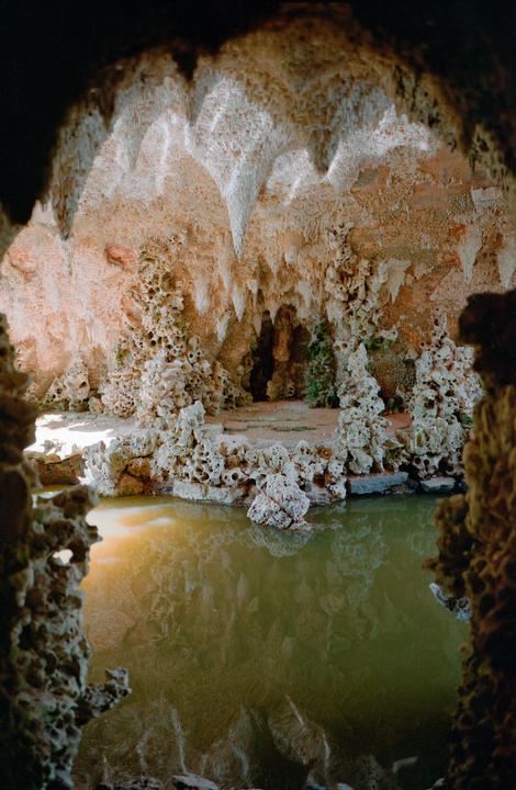 A cave with stalactites and stalagmites is illuminated by light from an opening, reflecting in a pool of water.