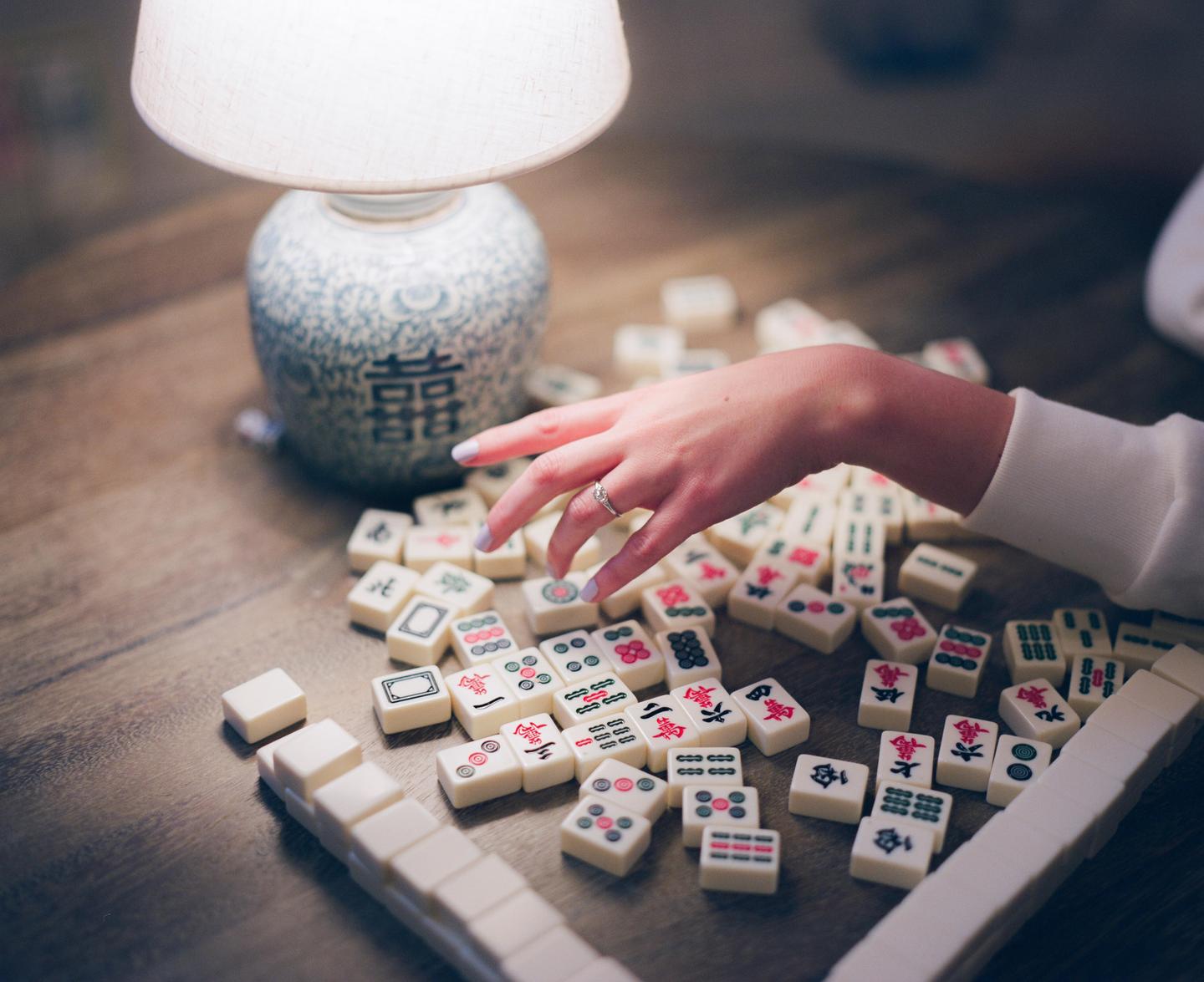 A hand reaches for a mahjong tile on a wooden table next to a lamp.
