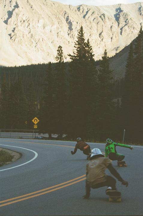 Three men on skateboards race down a winding mountain road lined with pine trees and a rocky mountain.