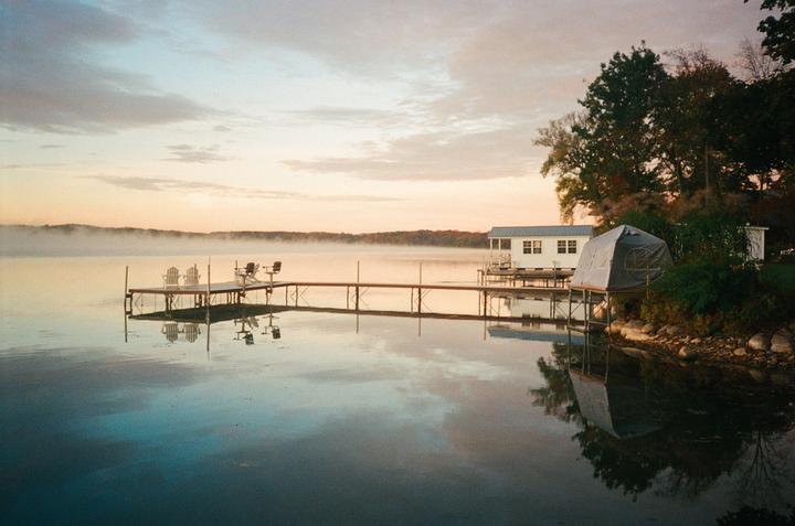 A long dock with chairs extends into a misty lake at sunrise, with a small house and tent on the shore.