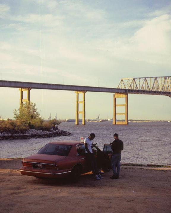Two men stand next to a red car parked on a dirt road next to a body of water with a bridge in the background.