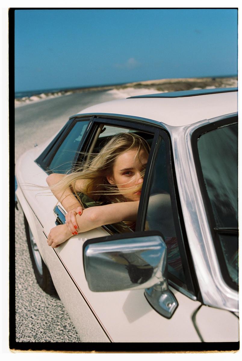 A woman with long blonde hair leans out of a car window on a sunny day.