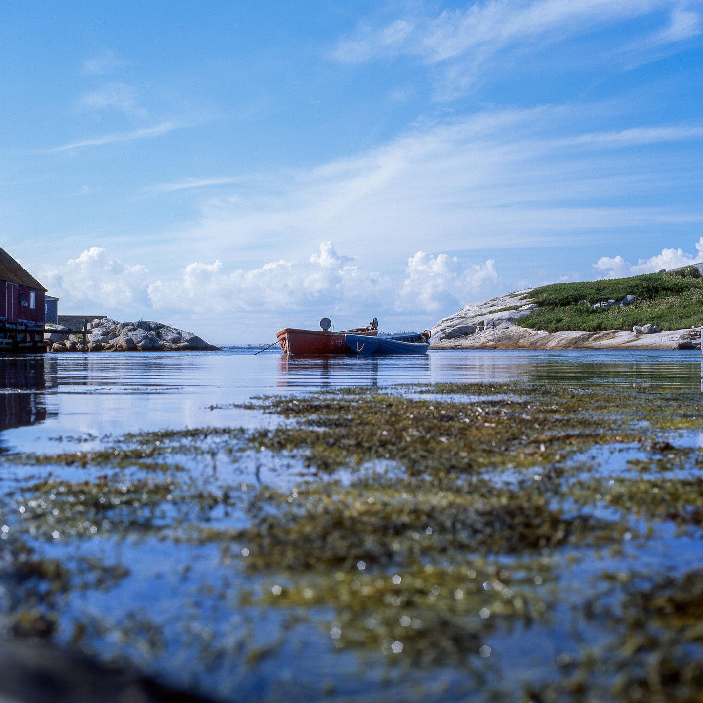 Two small boats float on calm water with a red building and rocky shore visible under a blue sky with clouds.