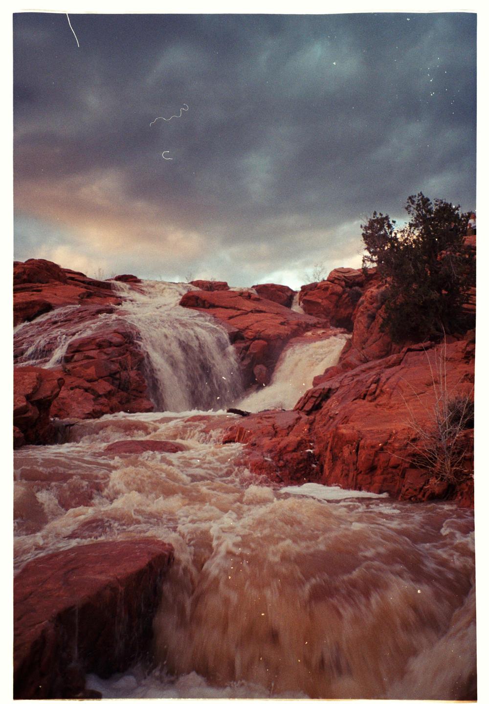 A waterfall cascades over red rock formations under a cloudy sky.