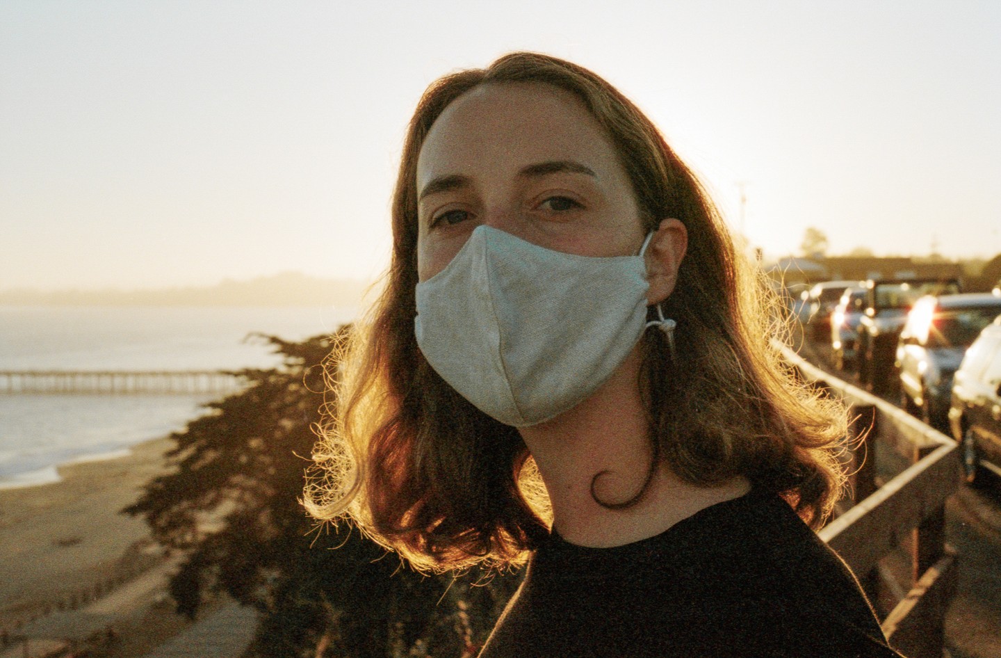 A woman wearing a face mask looks at the camera with a beach and pier in the background during golden hour.