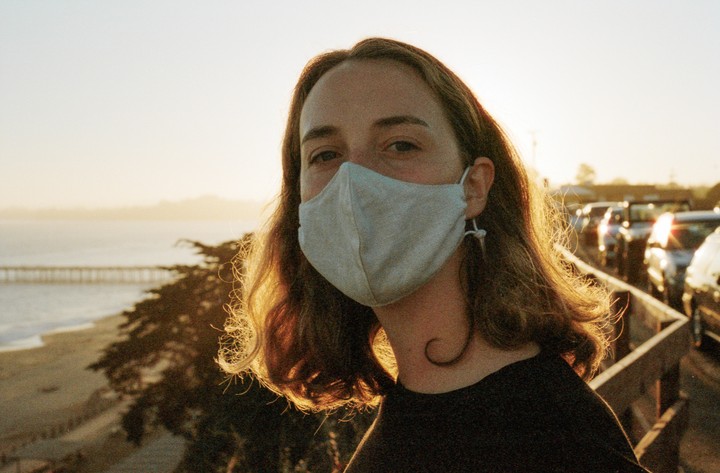 A woman wearing a face mask looks at the camera with a beach and pier in the background during golden hour.